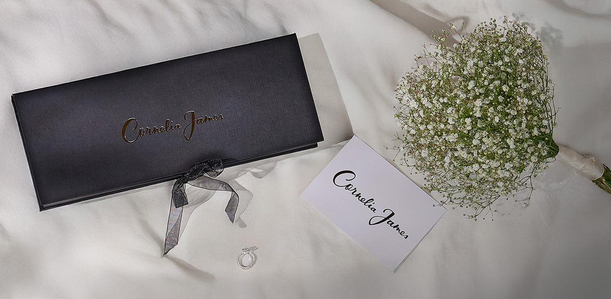 Black jewelry box with 'Carvela Kurt Geiger' branding, accompanied by a card and small bouquet of flowers on a light background.
