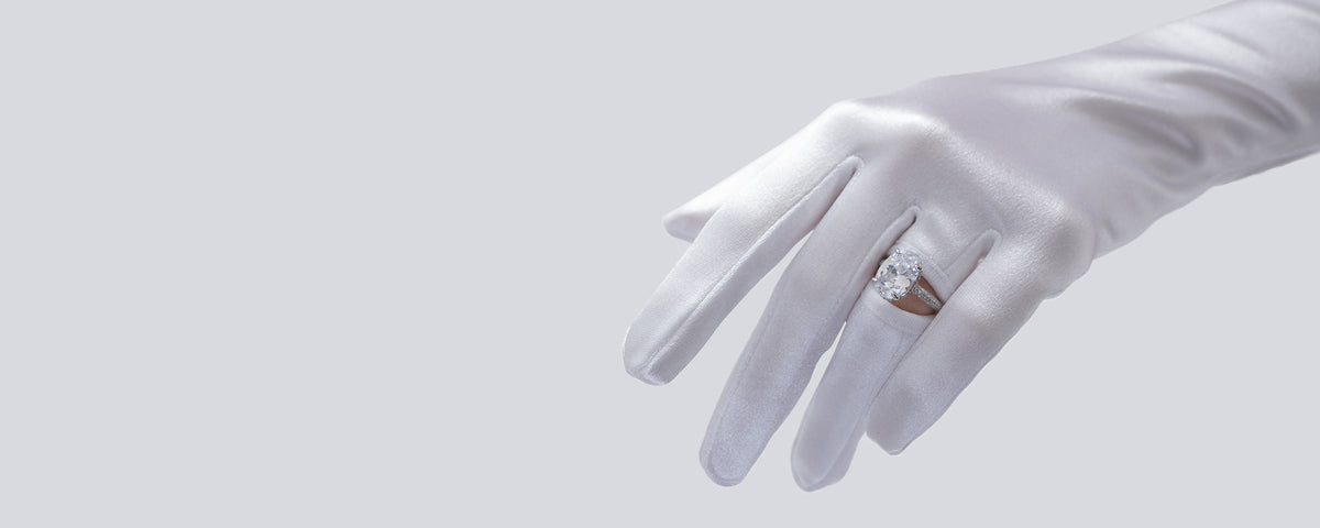 Hand wearing a white glove holding a ring with a diamond on a light gray background