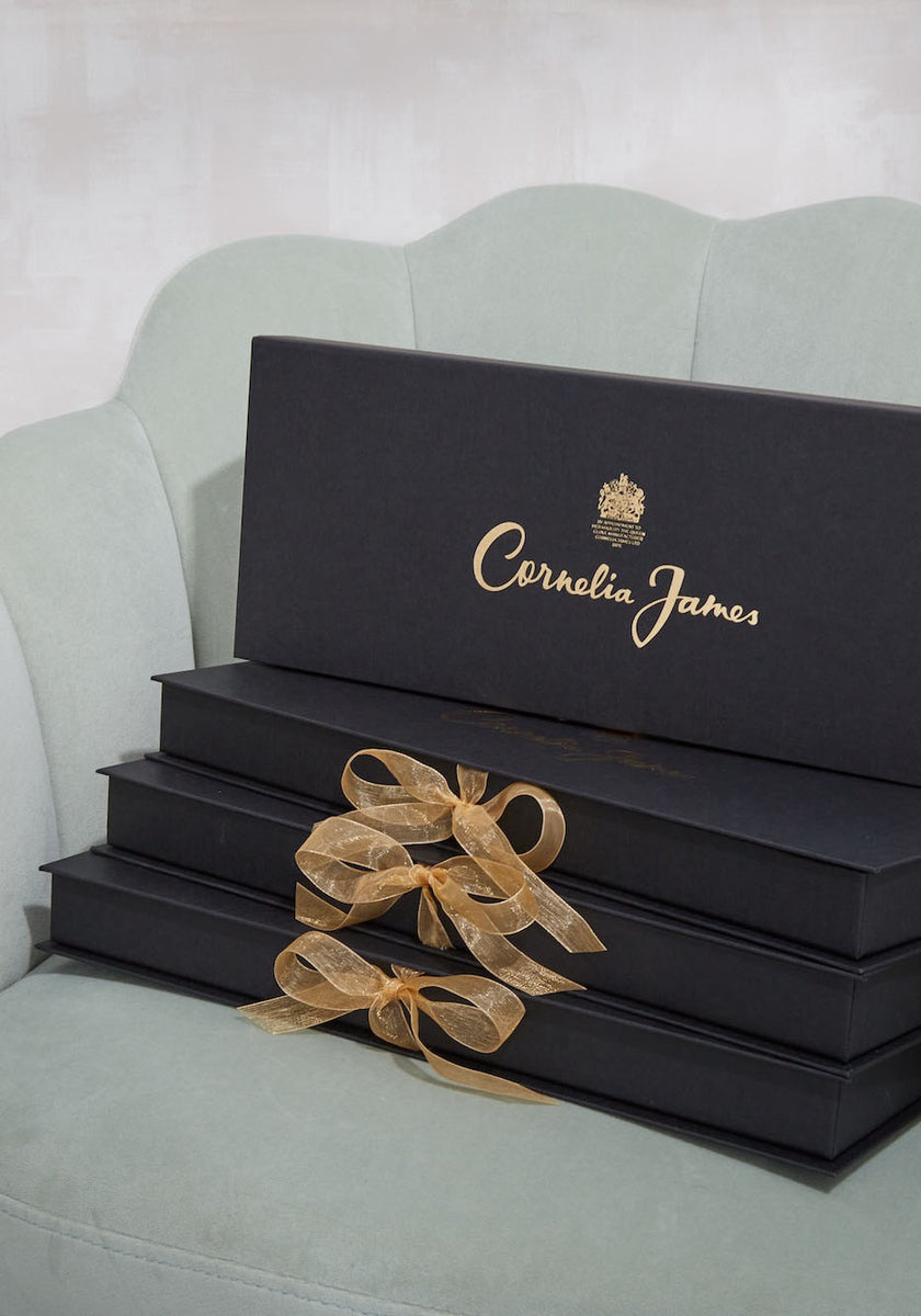 A stack of black rectangular gift boxes with gold 'Corkelia James' script and crest logo, tied with a sheer gold ribbon, displayed on a pale mint green upholstered chair.